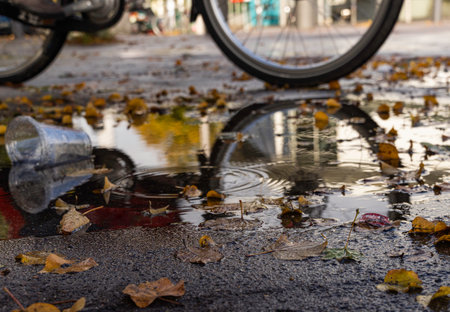 Puddle with colored autumn leaves and reflections in which a bicycle is passing and in which there is an empty transparent plastic cup, horizontalの写真素材