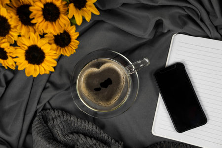 Heart-shaped glass cup filled with coffee on a glass saucer in front of gray satin fabric, a black smartphone on a lined notebook on the right also in the picture, horizontal, top viewの写真素材