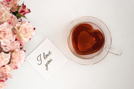 Heart-shaped glass teacup filled with red tea on a white background, light pink roses on the left and a white sticky note with the words I love you in the picture, top viewの写真素材