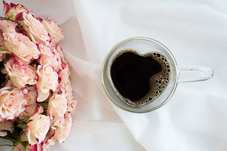Heart-shaped glass cup filled with coffee on white satin fabric, pink roses on left with pictured, top view, horizontalの写真素材