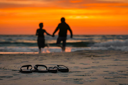 In the foreground are two pairs of flip flops on the beach, in the background the silhouettes of a woman and a man in the sunset as they walk hand in hand into the waterの写真素材