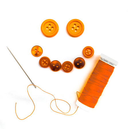 Orange buttons arranged to form a smiley face, sewing thread roll and sewing needle included in the picture, 1:1, white backgroundの写真素材