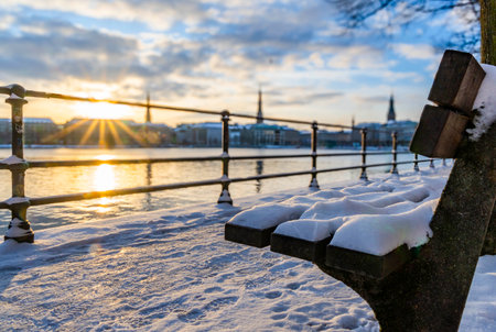 Snow-covered park bench at Ballindamm in Hamburg in the morning in winter at sunrise, the buildings on the Alster can be seen out of focus in the background, horizontalの写真素材