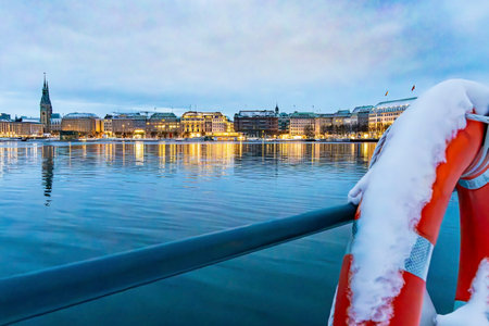 View from the LombardsbrÃ¼cke to the illuminated house facades on the Alster in Hamburg, part of a lifebuoy covered with snow can be seen in the foreground, horizontalの写真素材