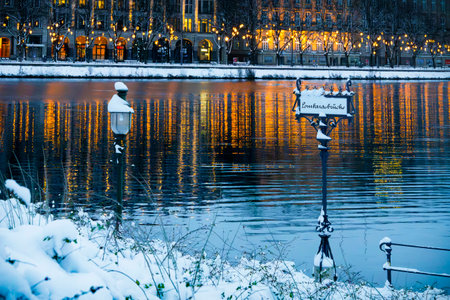 Vertical view of the Lombard Bridge sign covered in snow, in the background Christmas lights and illuminated house facades in Hamburg in winter, reflected in the water, horizontalの写真素材