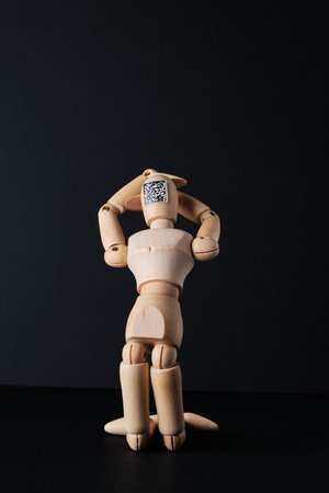 A wooden mannequin with a QR code is kneeling on a dark background. Selective focus.
