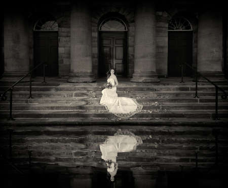 Beautiful Happy bride with wedding flowers bouquet sitting on stairs in front of a church looking out on water reflection  unusual bridal pictureの写真素材