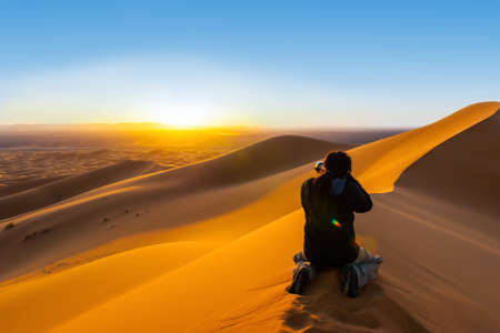 Handsome man with dreadlocks taking a photograph of sunset fron a sand dune の写真素材