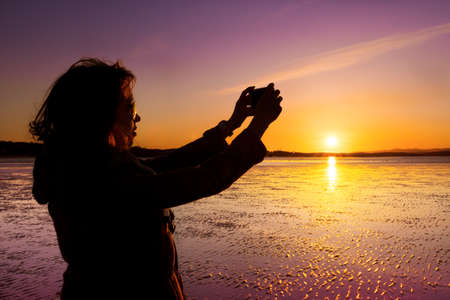 Beautiful young woman taking picture of herself, selfie, on a beach during sunset.の写真素材