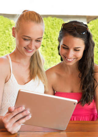 holidays, tourism, technology and internet - two beautiful girls looking at tablet pc computer in...