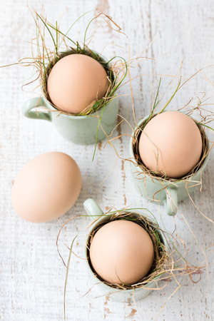 Uncolored natural easter eggs in espresso cups; happy easter concept; white wooden backgroundの写真素材