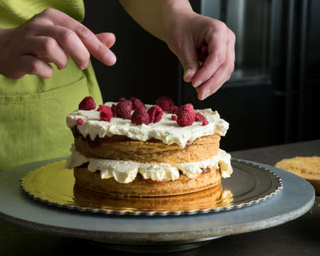 Woman decorating a delicious layered sponge cake with icing cream and raspberriesの写真素材