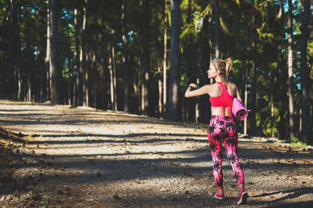 Young fit athletic woman in a forest wearing smart watch and holding yoga mat, walking away from the camera. Mind and body happiness conceptの写真素材