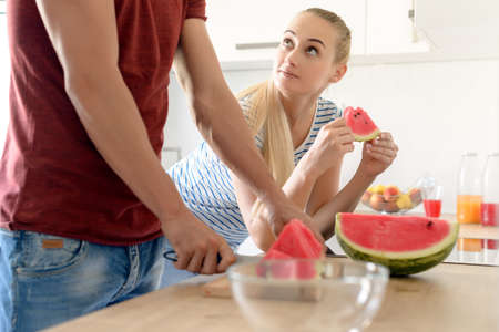 Young attractive woman in a kitchen eating red watermelon and looking at her husband, Couple in their large contemporary white kitchenの写真素材