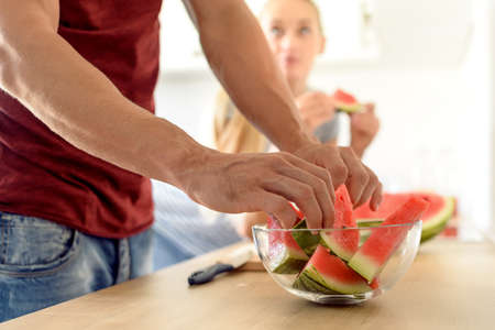 Young attractive woman in a kitchen eating red watermelon and looking at her husband, Couple in their large contemporary white kitchenの写真素材