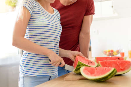 Young couple in a kitchen slicing red watermelon. Couple in their large contemporary white kitchenの写真素材