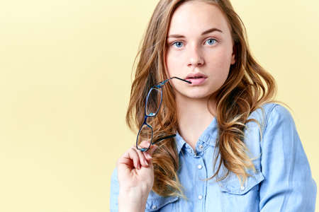 Beautiful teenager girl with ginger hair, freckles and blue eyes holding reading glasses, young woman with spectacles on yellow backgroundの写真素材