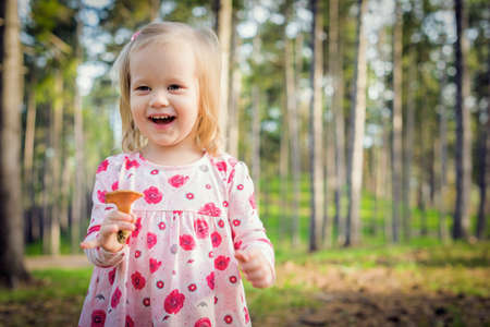 Cute blond toddler girl picking mushrooms in a forest. Happy child holding a mushroom and laughingの写真素材