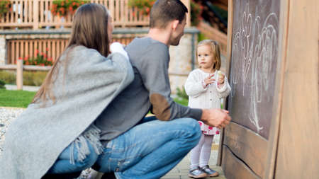 Young parents with their cute little girl drawing on blackboard. Family having fun outdoors, holding chalk and drawing.の写真素材