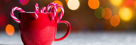 Red mug with candy canes, with christmas lights bokeh. Festive Christmas web banner with copy space
