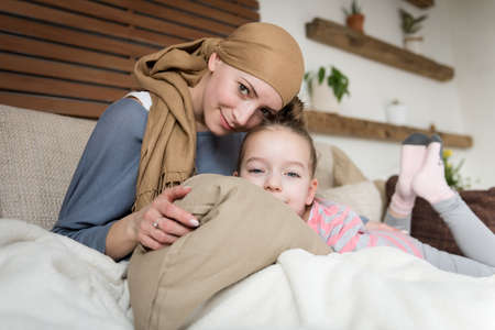 Young adult female cancer patient spending time with her daughter at home, relaxing on the couch. Cancer and family support concept.の写真素材