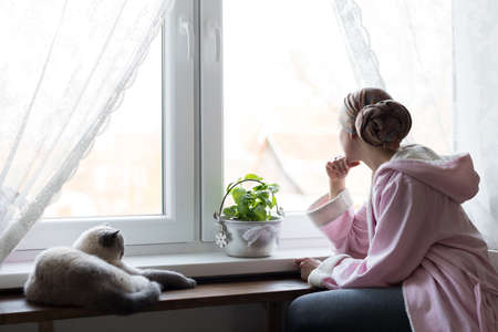 Young adult female cancer patient wearing headscarf and bathrobe sitting in the kitchen with her pet cat, looking out window.の写真素材