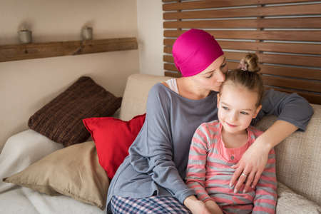 Young adult female cancer patient spending time with her daughter at home, relaxing on the couch. Cancer and family support concept.の写真素材