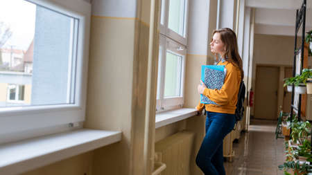 Young attractive female high school student standing by the window in the hallway at her school...