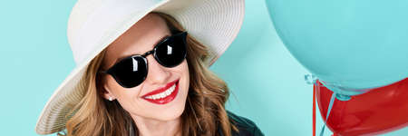 Beautiful woman in summer hat and sunglasses holding balloons. Attractive cool young woman fashion portrait over pastel blue banner background.の写真素材