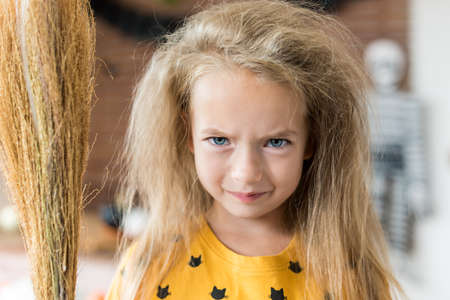 Cute little girl with messy hair, holding a broom and dressed up as a witch standing in Halloween decorated living room, looking at camera with angry facial expression. Halloween party concept.の写真素材