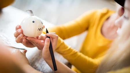 Cute young girl sitting at a table, decorating little white Halloween pumpkins with her mother. Hand...