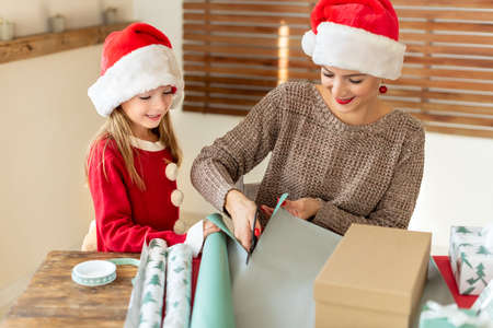 Mother and daughter wearing santa hats having fun wrapping christmas gifts together in living room. Candid family christmas time lifestyle background.の写真素材