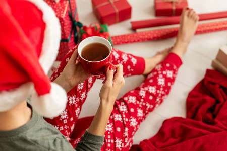 Young woman wearing santa hat and xmas pajamas sitting on the floor amongst wrapped christmas presents, drinking hot punch. Over the shoulder view.の写真素材