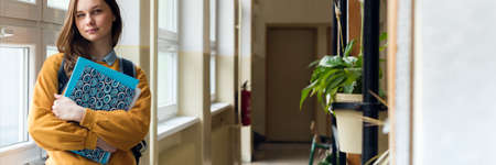 Young attractive female high school student standing by the window in the hallway at her school...