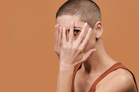 Beautiful young woman with shaved head covering her face with both hands and peeking through...
