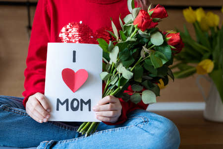 Unrecognizable young girl holding homemade greeting card and red roses for her mom. Family celebration concept. Happy Mother's Day or Birthday Backgroundの写真素材
