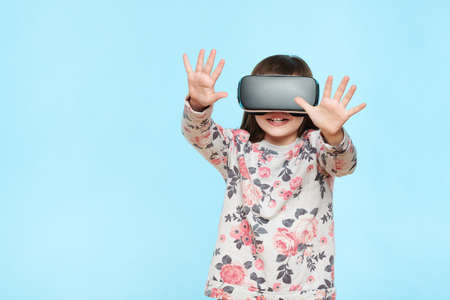 Studio portrait of cute young caucasian girl wearing virtual reality glasses against pastel blue background. Future gadgets technology concept.の写真素材