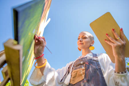 Beautiful young woman relaxing while painting an art canvas outdoors in her garden. Cancer survivor,...