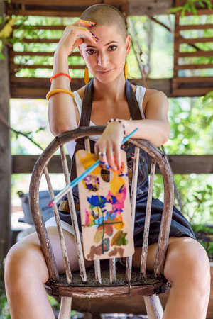 Playful portrait of a young gorgeous female painter artist looking and smiling at camera, sitting on an old wooden chair, holding palette and a paintbrush.の写真素材