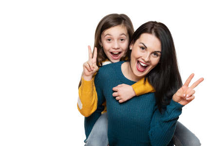 Beautiful young mother and her daughter making peace sign hand gesture, laughing and looking at camera. Isolated over white background. Family love connection concept.の写真素材