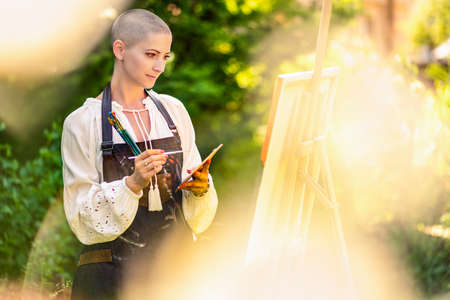 Beautiful young woman relaxing while painting an art canvas outdoors in her garden. Cancer survivor,...