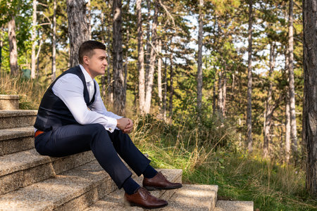 Handsome young groom wearing elegant and stylish dark blue suit outdoors portrait. Stylish groom sitting on stairs side view.の写真素材