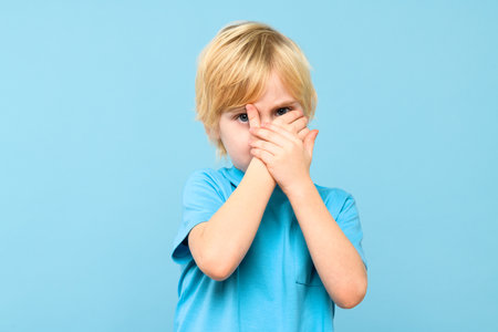 Emotional young boy covering face with hands isolated over pastel blue background. Mental wellbeing and safety in childhood age.の写真素材