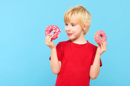 Cute little boy with blond hair and freckles having fun with glazed donuts, sticking tongue out. Children and sugary junk food concept.の写真素材