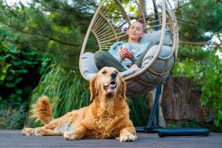Young female cancer patient relaxing in hanging chair on the patio with her emotional support therapy dog lying by her side.の写真素材