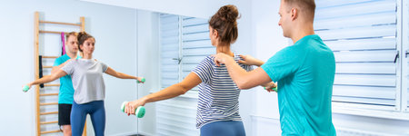 Woman with shoulder pain in physical therapy. Physical therapist exercising with female patient doing shoulder mobility exercises using a lightweight dumbbells.の写真素材