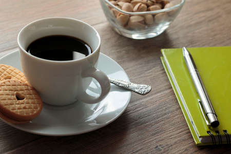 Cup of coffee with saucer on wooden background with partial view of a notebook and pistachio bowlの写真素材