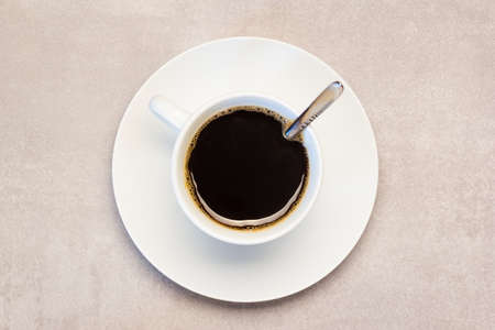 The handle of the cup and the spoon are arranged to form a checkmark. The white cup is filled with dark fresh coffee. During the roasting process the characteristic flavor of coffee arises. The cup is on a countertop ready to be served. Image can fit menu cards.の写真素材