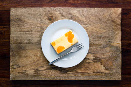 A piece of cheesecake with tangerine slices on a plate with fork on dark wooden background