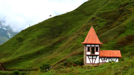 A small house with German architecture, open windows, white walls with brown stripes, and an orange roof. Located on the mountain of green grass, low trees, narrow trails and behind the cloudy sky.の写真素材
